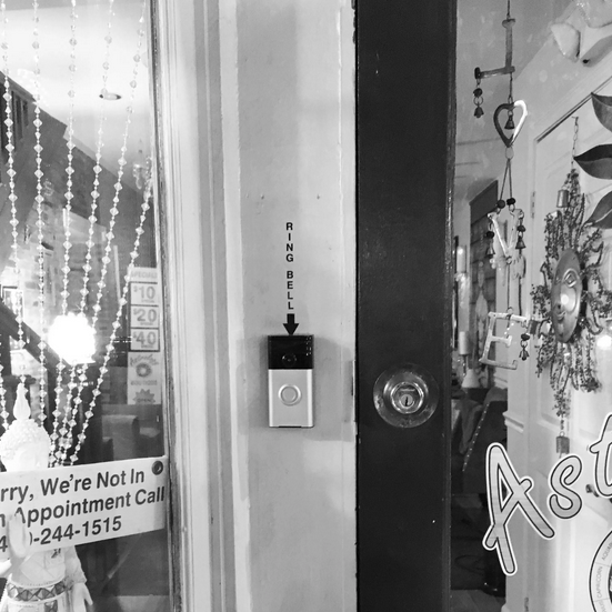 Black and white photo of a the door to an astrology shop with a Ring doorbell in the center and with sticky letters on the door frame above the bell, reading vertically "RING BELL" with an arrow pointing to the camera. In the window, a beaded curtain and statue of the Buddha. Service rates of $10, $20, and $40 are listed and a sign that says, "Sorry, we're not in for appointment call [Baltimore phone number partially obscured]. In the glass door, a sun-shaped wind chime and mobile spelling LOVE hang.