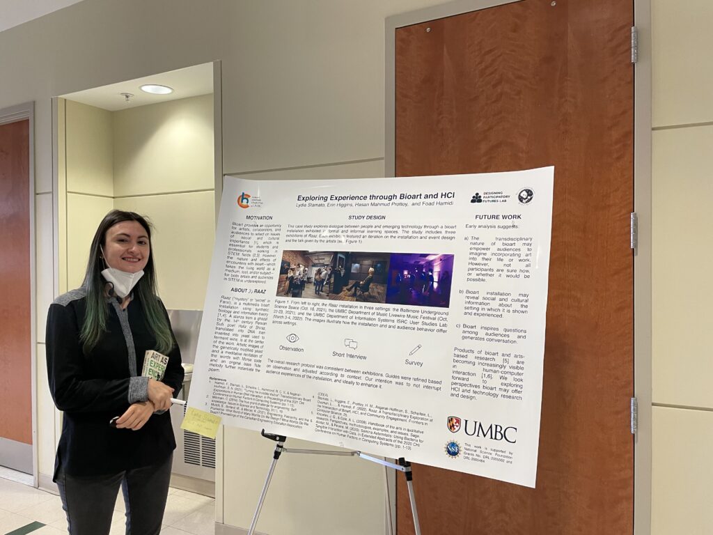 Lydia standing to the left of her poster, "Exploring Experience through Bioart and HCI" which is on a poster stand in a hallway. Lydia is holding a book, the title visible, "Art as Experience" by John Dewey. She is also holding a pen and half eaten oreo and wearing a kn95 mask pulled down on her chin to show a big smile.